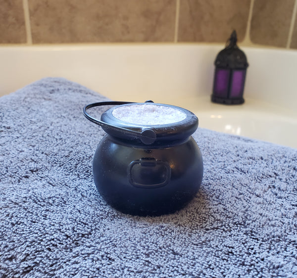 A black plastic cauldron filled with bath bomb ingredients. The cauldron sits on a gray towel with a bathtub and purple and black lantern in the background.