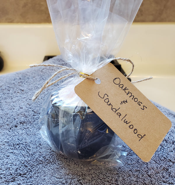 A packaged cauldron bomb wrapped in cellophane with a handwritten kraft label reading "Oakmoss & Sandalwood". The bath bomb sits on a gray towel over a bathtub.