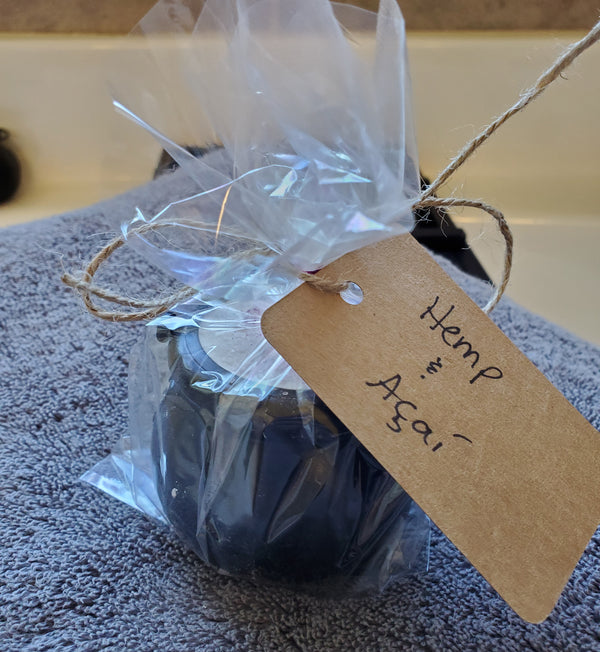 A packaged cauldron bomb wrapped in cellophane with a handwritten kraft label reading "Hemp & Acai". The bath bomb sits on a gray towel over a bathtub.