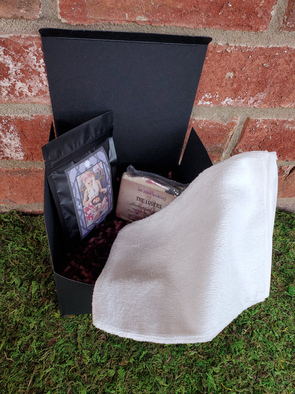 A black cardboard box containing purple shredded paper, one bar of The Lovers soap, one white bath mitt, and one bag of The Lovers tea. The box is sitting on a bed of moss against a red brick wall.