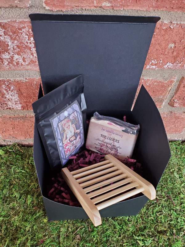 A black cardboard box containing purple shredded paper, one bar of The Lovers soap, one wooden soap ladder, and one bag of The Lovers tea. The box is sitting on a bed of moss against a red brick wall.