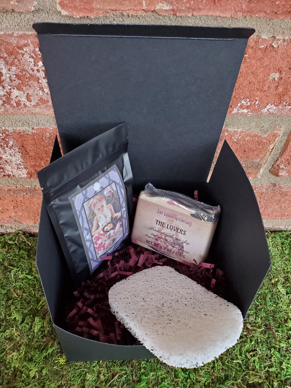 A black cardboard box containing purple shredded paper, one bar of The Lovers soap, one antibacterial soap saver pad, and one bag of The Lovers tea. The box is sitting on a bed of moss against a red brick wall.