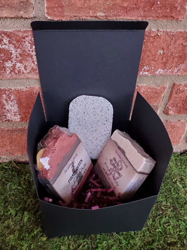 A black cardboard box containing purple shredded paper, two random soap bars, and one antibacterial soap saver pad. The box is sitting on a bed of moss against a red brick wall.