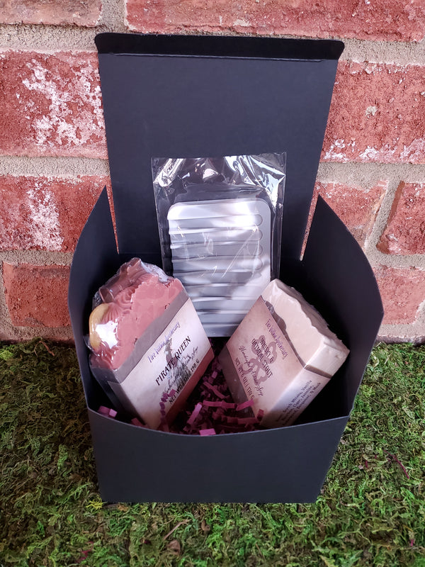 A black cardboard box containing purple shredded paper, two random soap bars, and one silicone soap saver. The box is sitting on a bed of moss against a red brick wall.