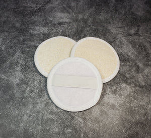 Three round luffa pads, two facing forward and one facing backward. The front is made of natural luffa, and the back is made of cottony fabric and has an elastic band for easy holding. There is a gray background.