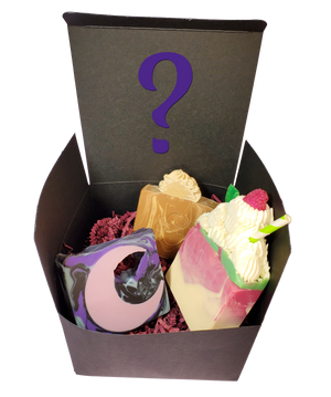 A black cardboard box containing purple shredded paper and three random soap bars. There is a purple question mark on the box.