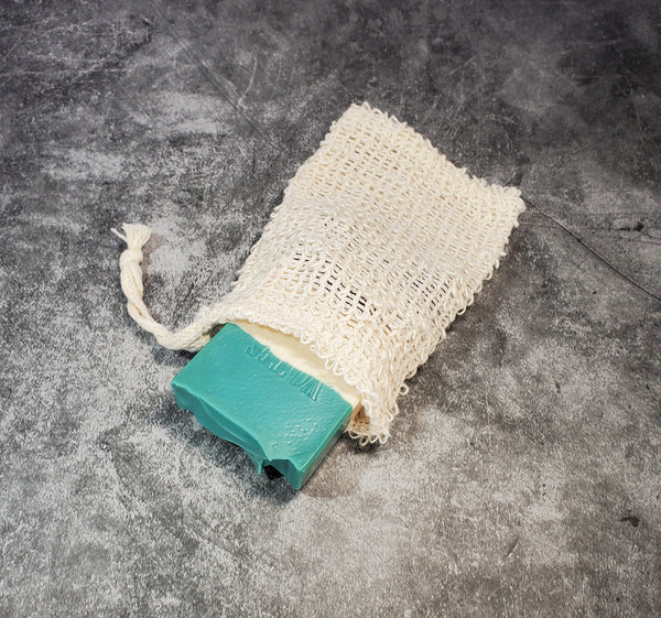 A sisal soap saver pouch, made of natural, beige fibers and a drawstring opening. The pouch contains a soap bar to demonstrate size. The pouch is on a gray background.