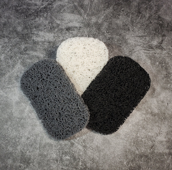 Three antibacterial soap saver pads, one gray, one white, and one black, against a gray background. Each pad is an oblong shape made of tight plastic squiggles to allow air circulation and water drainage to keep the soap dry between uses.
