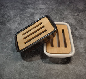 Two rectangular, ceramic soap dishes, one dark brown and one white, each accompanied by a bamboo slotted tray. The background is gray.