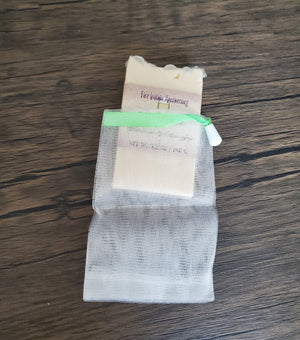 One mesh soap saver pouch with a tall and skinny soap bar tucked inside it to demonstrate size and use. The soap saver pouch is made of thin, white, tightly woven nylon netting and is tied with a green ribbon. The background is dark wood.