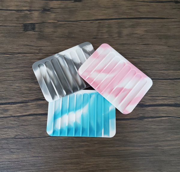 Three silicone soap savers, one gray and white, one pink and white, and one aqua blue and white, against a dark wood background. The soap savers are oblong silicone pads with grooves to allow for water drainage to keep the soap dry between uses.