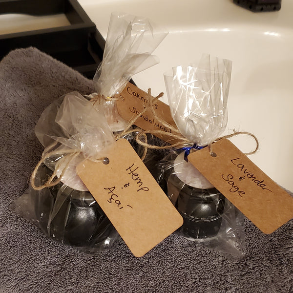 Three packaged cauldron bath bombs sitting on a gray towel on a tray above a bathtub. Each bath bomb is wrapped in clear cellophane, tied with twine, and labeled with a handwritten kraft label depicting each of the three available scents.