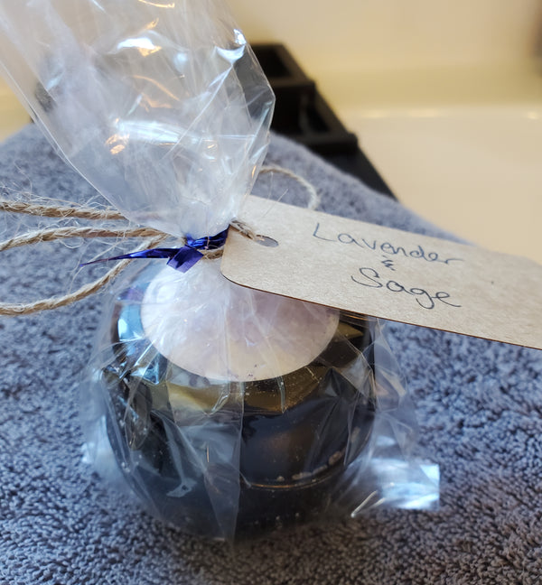 A packaged cauldron bomb wrapped in cellophane with a handwritten kraft label reading "Lavender & Sage". The bath bomb sits on a gray towel over a bathtub.