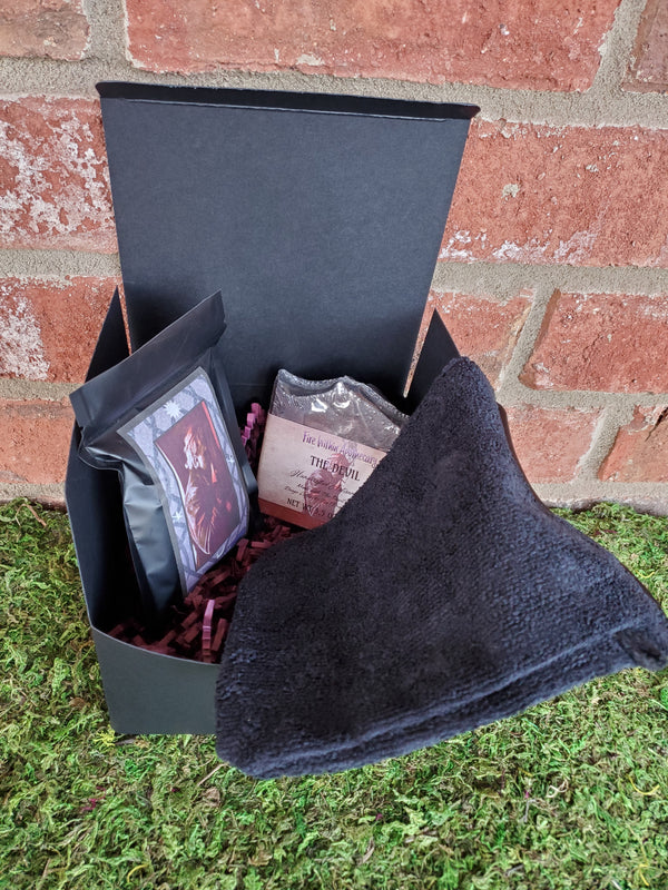 A black cardboard box containing purple shredded paper, one bar of The Devil soap, one black bath mitt, and one bag of The Devil tea. The box is sitting on a bed of moss against a red brick wall.