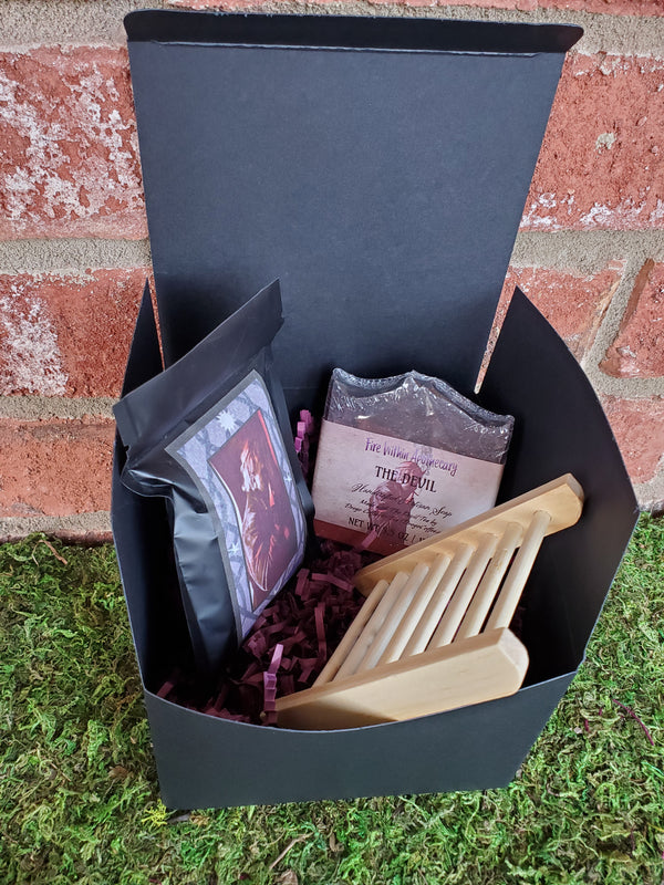 A black cardboard box containing purple shredded paper, one bar of The Devil soap, one wooden soap ladder, and one bag of The Devil tea. The box is sitting on a bed of moss against a red brick wall.