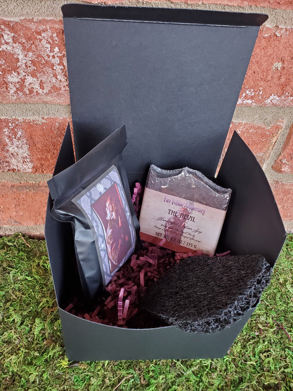 A black cardboard box containing purple shredded paper, one bar of The Devil soap, one antibacterial soap saver pad, and one bag of The Devil tea. The box is sitting on a bed of moss against a red brick wall.