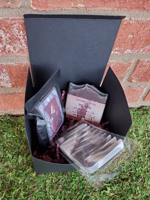 A black cardboard box containing purple shredded paper, one bar of The Devil soap, one silicone soap saver, and one bag of The Devil tea. The box is sitting on a bed of moss against a red brick wall.