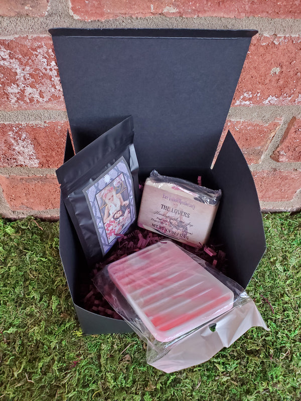 A black cardboard box containing purple shredded paper, one bar of The Lovers soap, one silicone soap saver, and one bag of The Lovers tea. The box is sitting on a bed of moss against a red brick wall.