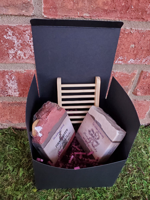 A black cardboard box containing purple shredded paper, two random soap bars, and one wooden soap ladder. The box is sitting on a bed of moss against a red brick wall.