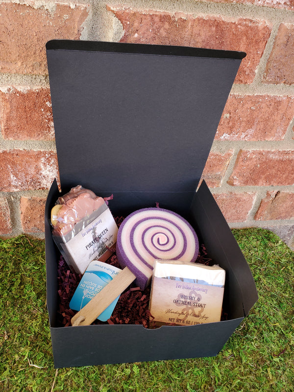 A black cardboard box containing purple shredded paper, two random soap bars, and one lollipop sponge. The box is sitting on a bed of moss against a red brick wall.