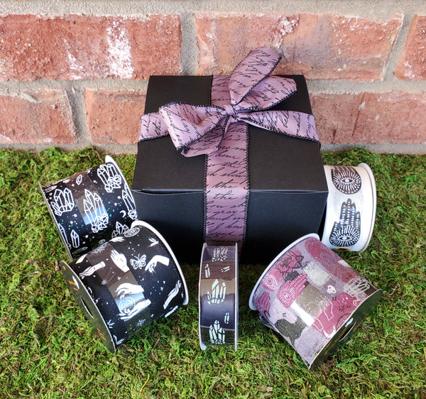 A black cardboard box wrapped in purple ribbon with illegible black writing on it. The ribbon is tied in a bow. Five spools of ribbon surround the box, each in varying patterns to represent a random and incomplete sample of ribbons used for the gift boxes. The box is sitting on a bed of moss against a red brick wall.