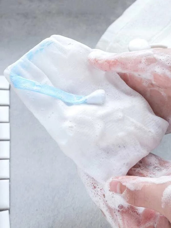 One mesh soap saver pouch covered in soap suds, held by two sudsy hands, to demonstrate use. One small soap bar is inside the pouch. The pouch is made of thin, white, nylon netting and is tied with light blue ribbon.