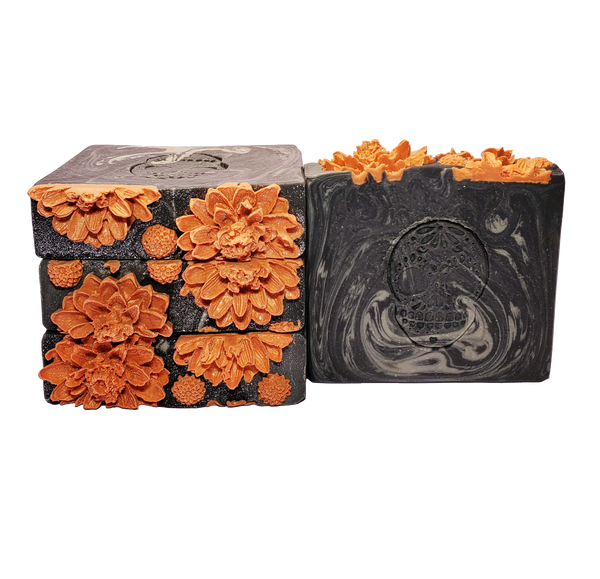 Four bars of Death soap. The bars are black with gray swirls, with a sugar skull stamped onto the bar face. The soaps are topped with orange soap flowers and glitter.