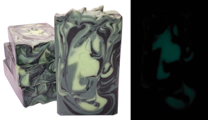 One bar of Medusa soap shown both in the light and in the dark. The bar contains swirls of light green, shimmery dark green, and black. A design of two entwined snakes is stamped on each bar. The photo of the soap bar in the dark demonstrates that the light green swirls glow in the dark.