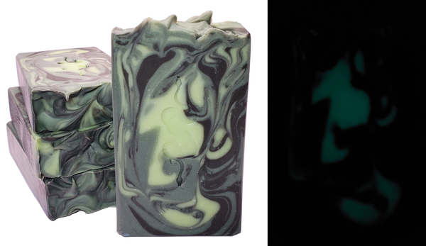 One bar of Medusa soap shown both in the light and in the dark. The bar contains swirls of light green, shimmery dark green, and black. A design of two entwined snakes is stamped on each bar. The photo of the soap bar in the dark demonstrates that the light green swirls glow in the dark.