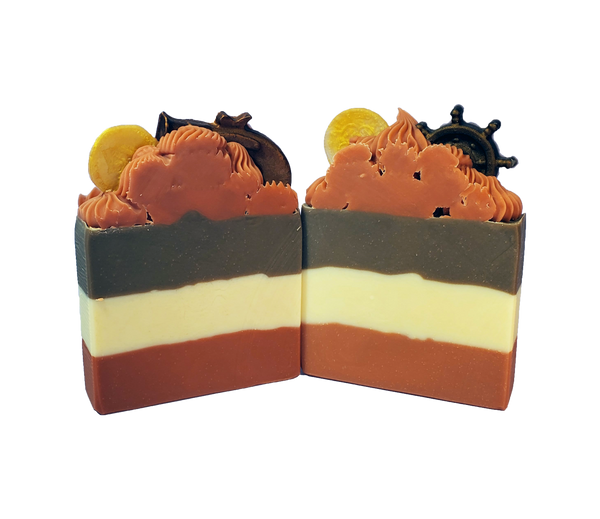 Two bars of Pirate Queen soap. Each bar is layered, with dark orange at the bottom, creamy white in the middle, and dark brown on top. The bars feature a dark orange piped top with small soaps in the shape of gold doubloons and either a coppery pistol or a dark brown ship wheel.